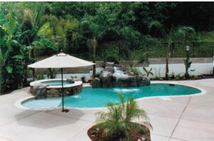 An inviting backyard swimming pool with a cascading stone waterfall feature and integrated hot tub. The pool area is completed with a patterned concrete deck, a large patio umbrella for shade, and tropical landscaping, including palm trees and lush greenery, behind a black wrought iron fence.